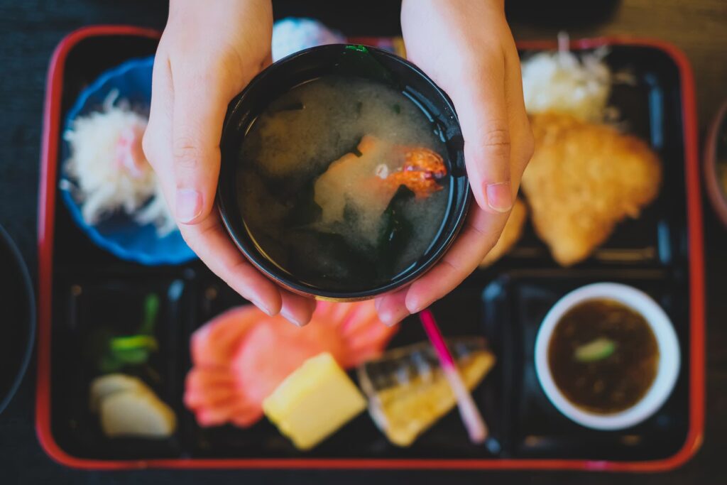 A person holding a black bowl of traditional Japanese miso soup over a bento meal tray.