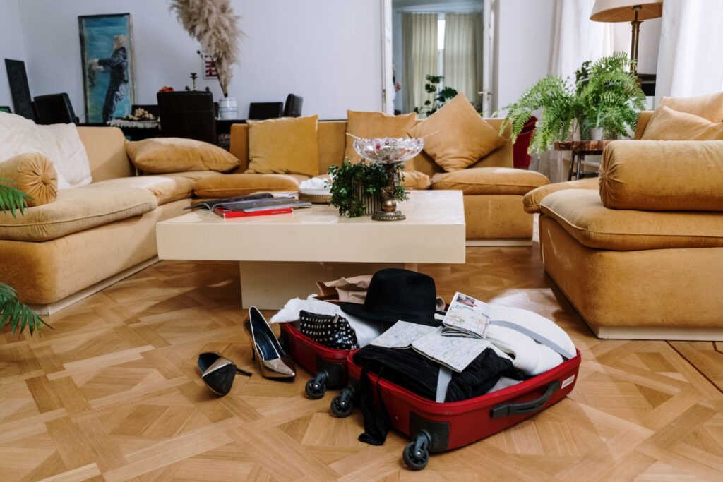 An overpacked red suitcase overflowing with clothes, a black hat, and shoes scattered on a wooden living room floor.