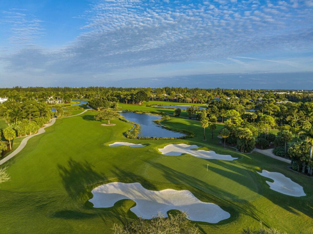 An aerial view of a manicured green golf course with sand traps, water hazards, and trees under a partly cloudy sky.
