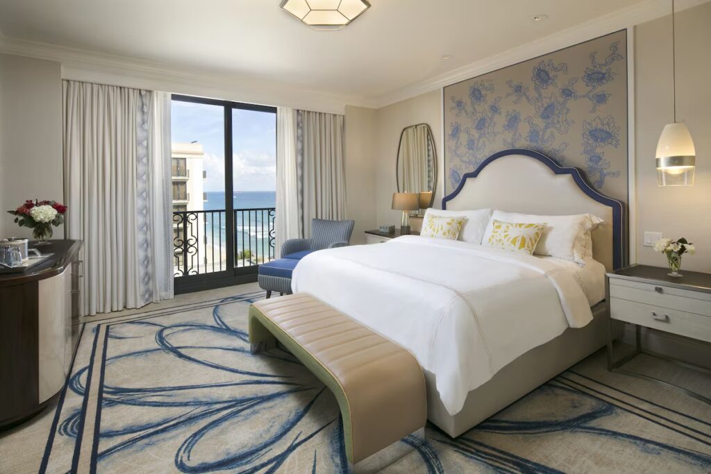 A bright, luxurious hotel bedroom with a large bed, floral headboard pattern, and an open balcony overlooking the ocean.