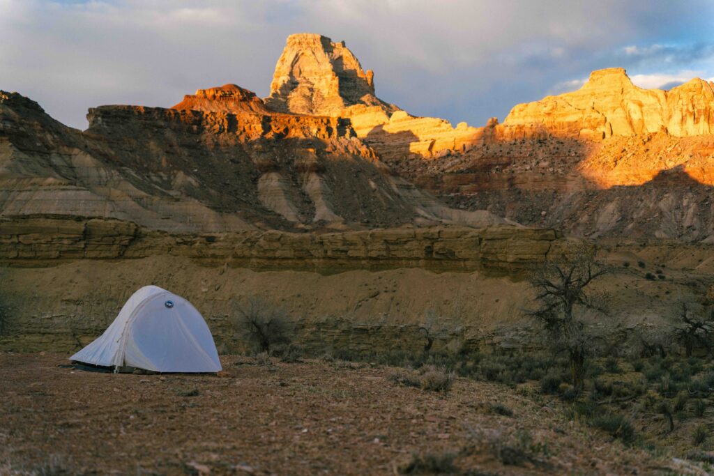 A solitary tent sits in a rugged canyon landscape with a massive stone peak glowing orange under the setting sun.