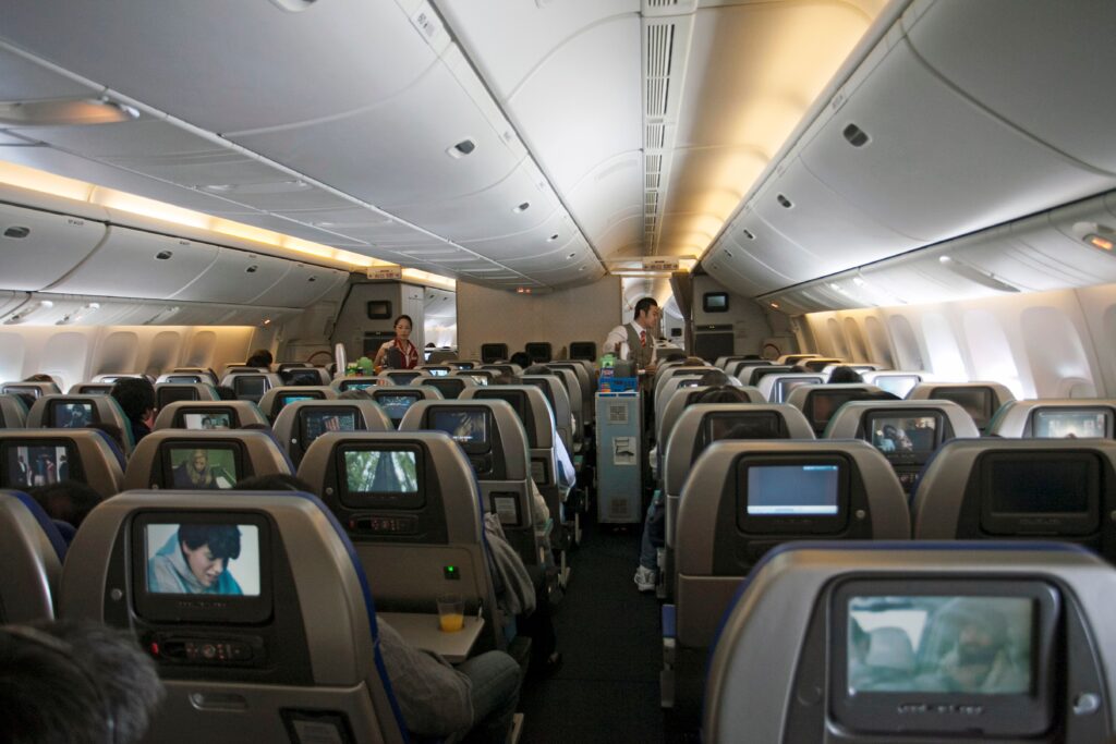 Interior view of a crowded passenger airplane cabin with rows of economy seats and individual screens.