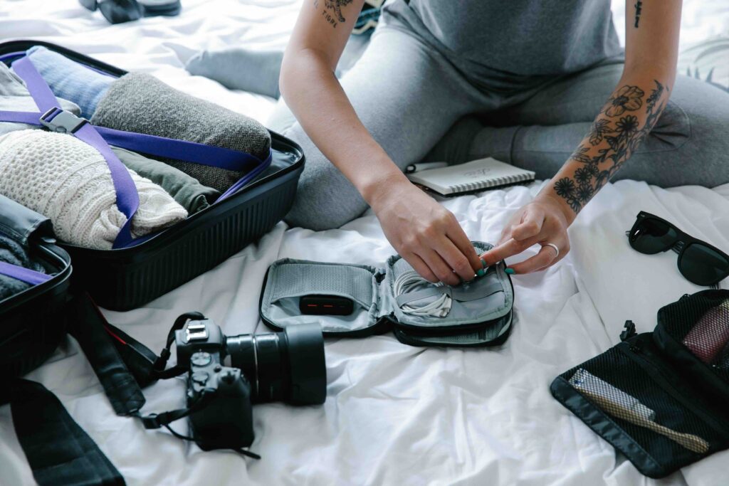 Woman sitting on a bed packing charging cables and a power bank into a small travel electronics organizer pouch.