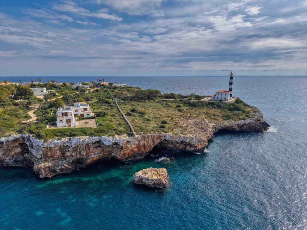A striped lighthouse stands on a rugged cliff over clear Mediterranean waters.