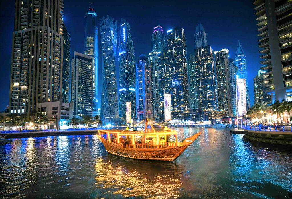 Illuminated wooden dhow boat cruising past glowing skyscraper towers, a popular must see in Dubai.