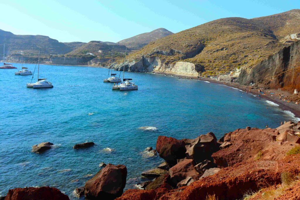 Catamarans floating near red volcanic cliffs on Greece island beaches.