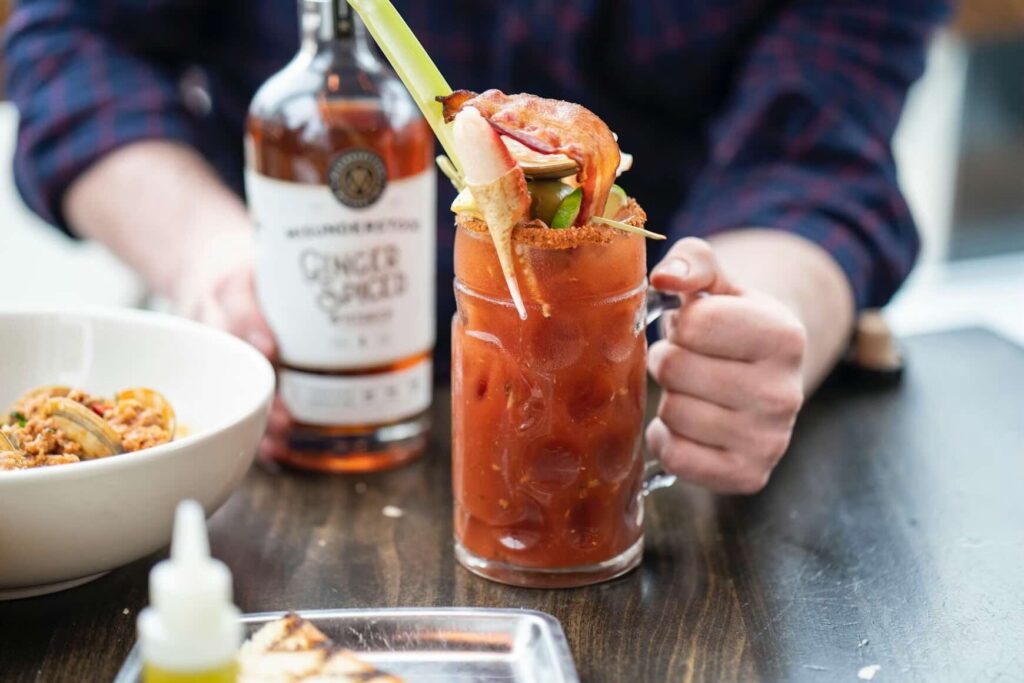 A person holding a large glass mug of a Bloody Mary topped with shrimp and bacon. Source: Unsplash