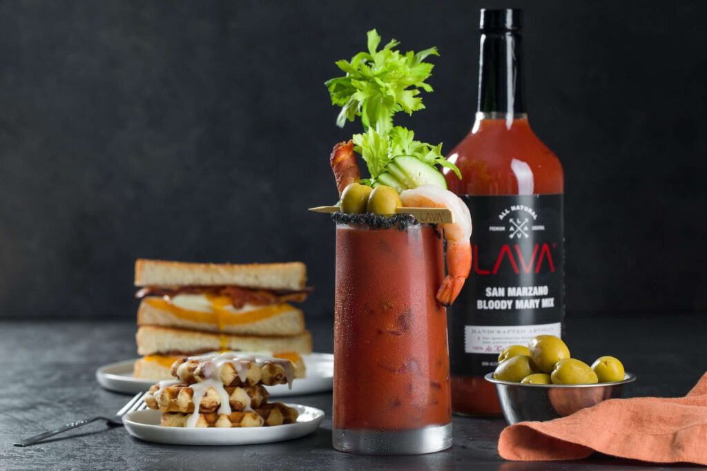A tall Bloody Mary heavily garnished with shrimp, bacon, and celery next to a grilled cheese sandwich. Source: Pexels