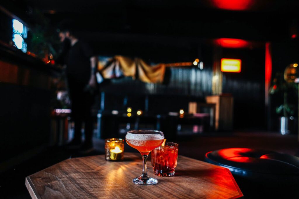 Two brightly colored cocktails sit on a low wooden table in a dimly lit bar lounge. - Source: Pexels
