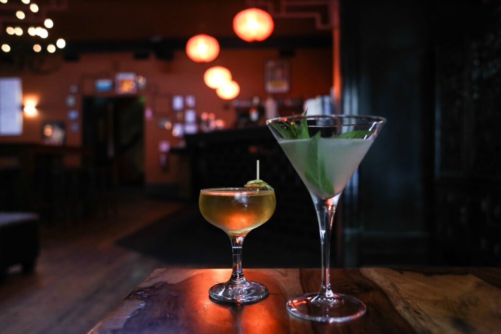 Two cocktails on a wooden table in a bar with glowing round lanterns in the background. - Source: Pexels